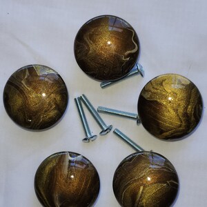 May include: Five round, brown and gold cabinet knobs with white swirls. The knobs have a shiny, metallic finish. The knobs are arranged in a circle with screws in the center.