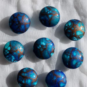 May include: Eight round, blue and turquoise decorative beads with a marbled effect and copper-colored veining. The beads are arranged on a white, textured surface. The beads have a glossy finish.