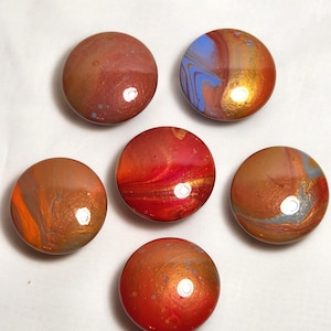 May include: A set of five circular cabinet knobs featuring a glossy, multi-colored pour paint finish. Each knob displays a unique swirl of warm tones including red, orange, gold, and copper, with some accents of blue and grey throughout the surfaces.