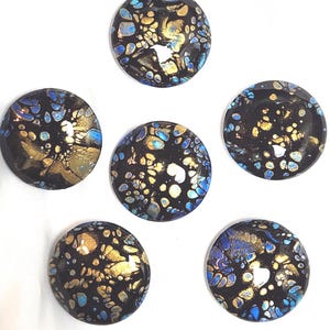May include: Six circular cabochons featuring a black base with an abstract fluid paint design in metallic gold and bright blue. Each piece displays a glossy, smooth finish with scattered, organic cell-like patterns across the surface.