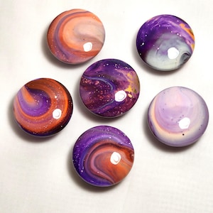 May include: Five circular cabinet knobs featuring a glossy, poured paint finish with swirling patterns in shades of purple, orange, white, and deep violet. Each knob displays a unique marbled effect with small white speckles and reflective highlights.