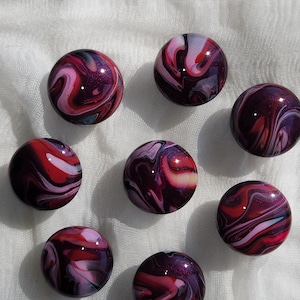 May include: Eight decorative glass marbles with swirling patterns in shades of red, purple, and white. The marbles have a glossy finish and are arranged on a white fabric surface.