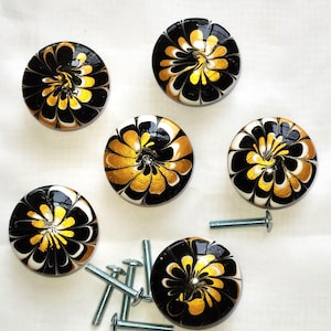 May include: A set of six round cabinet knobs featuring a swirling black, white, and metallic gold floral pattern. Each knob has a smooth, glossy finish and comes with a silver-toned metal mounting screw for installation on furniture or cabinetry.