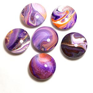 May include: Six circular cabochons featuring a glossy finish with fluid acrylic paint pours in shades of purple, orange, white, and dark brown. Each piece displays unique marbled patterns with swirling colors and small paint droplets across the surface.