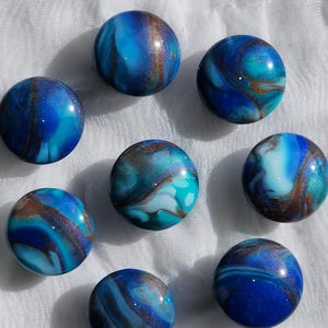 May include: Eight round, marbled glass beads in shades of blue, teal, and white with bronze-colored accents. The beads have a glossy finish and are arranged on a white, textured surface. The overall effect is a swirling, oceanic pattern.