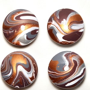 May include: Four circular buttons featuring a marbled pattern with swirls of metallic copper, dark brown, white, and grey. Each button has a smooth, glossy finish and a rounded surface, displaying a unique fluid paint design across the top.