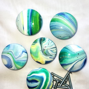 May include: Six round cabinet knobs featuring a marbled paint design in shades of blue, green, white, and yellow. Each knob has a smooth, glossy finish with swirling patterns. Several metal mounting screws are included with the hardware set.