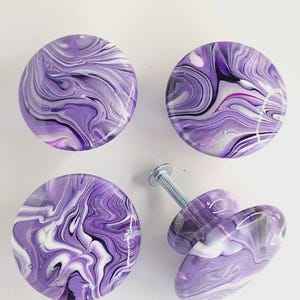 May include: Four round cabinet knobs featuring a glossy, marbled finish with swirling patterns of purple, white, and black. Each knob has a smooth, domed surface. One knob is positioned on its side, revealing a silver metal screw attachment.
