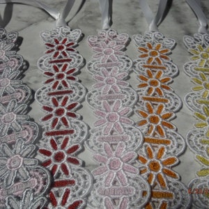 May include: Five white lace bookmarks with different colored floral designs. The bookmarks are made of white lace with a ribbon attached to the top. The floral designs are in shades of pink, red, orange, and yellow.