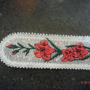 以下が含まれることがあります： 白いレースのしおりに、緑の茎と赤い花が刺繍されています。しおりはギザギザの縁をしています。