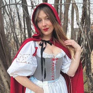 May include: A red hooded cape and a white and blue striped dress. The outfit includes a gray corset with red and blue lacing, a white blouse with lace sleeves, and a white pouch with blue embroidery. The outfit is reminiscent of a fairytale.