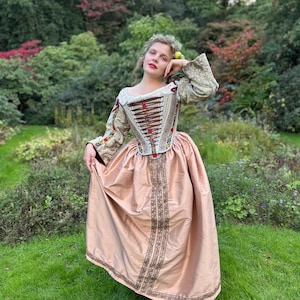 May include: A historical costume featuring a cream-colored bodice with brown and gold accents, and a pink skirt. The sleeves are gold with floral embroidery. The outfit is worn outdoors in a garden setting.