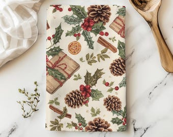 May include: A rectangular cream-colored kitchen towel with a festive holiday pattern. The design includes illustrations of pine cones, holly leaves, red berries, wrapped gifts, cinnamon sticks, and orange slices. A wooden spoon and a striped cloth are in the background.