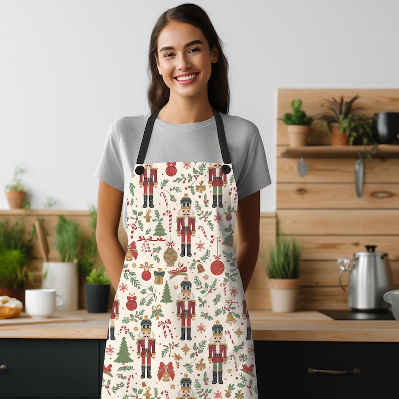 May include: A kitchen apron featuring a repeating pattern of nutcrackers, red Christmas ornaments, green pine trees, candy canes, and holly leaves on a cream background. The apron has black neck and waist straps and a straight top edge design.