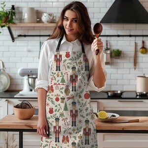 May include: A kitchen apron featuring a repeating pattern of nutcrackers, red and gold Christmas ornaments, green pine trees, candy canes, and bows on a cream background. The apron has black neck and waist straps for secure fastening.