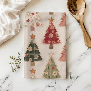 May include: A folded beige Christmas tea towel featuring appliqued Christmas trees in red, green, and pink floral patterns. Each tree is topped with a gold star. A wooden spoon and a striped cloth are in the background.