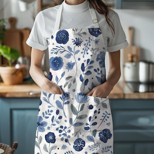 Op de afbeelding: Een wit schort met een blauw bloemenpatroon van diverse bloemen, bladeren en stengels. Het schort is voorzien van een witte nekband met een verstelbare metalen gesp en twee voorzakken op heuphoogte voor extra opbergruimte en gebruik.