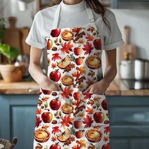 May include: A white apron featuring a repeating pattern of red apples, apple pies, cinnamon sticks, and colourful autumn leaves. The apron includes two front pockets and white neck and waist ties, providing full coverage for the torso and upper legs.
