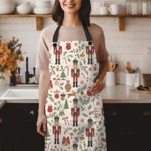 May include: A cream-colored apron featuring a repeating pattern of red and green nutcrackers, Christmas ornaments, pine trees, candy canes, and holly leaves. The apron has black neck straps and black buttons at the top corners of the bib section.