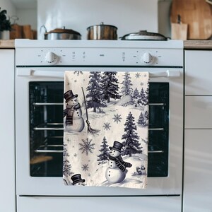 以下が含まれることがあります： グレーと白の色合いで雪だるま、松の木、雪の結晶が描かれた冬のシーンのキッチンタオル。タオルはオーブンのドアに掛けられています。
