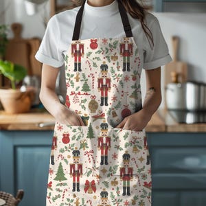 May include: A cream-colored apron with a festive Christmas pattern featuring nutcrackers, ornaments, candy canes, and Christmas trees. The apron has a black neck strap and two front pockets. The apron is worn by a person.