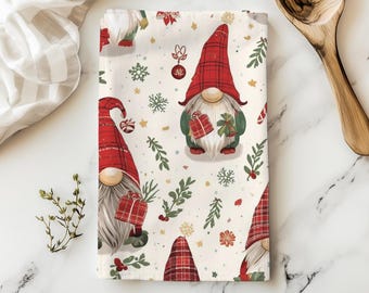 May include: A cream-colored Christmas-themed kitchen towel featuring a repeating pattern of gnomes with red plaid hats, holding gifts. The towel also has snowflakes, sprigs, and ornaments. A wooden spoon and a striped cloth are in the background.