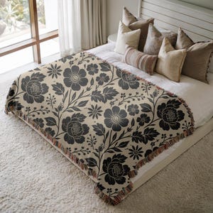 May include: A woven throw blanket featuring a dark charcoal gray floral pattern on a light beige background. The blanket has a decorative fringe along the edges and is draped over a bed with several neutral-toned pillows and white bedding.