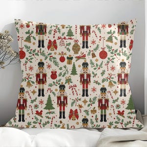 May include: A decorative pillow with a festive Christmas pattern. The design features repeating images of nutcrackers in red and black uniforms, along with Christmas trees, ornaments, and candy canes on a cream background.