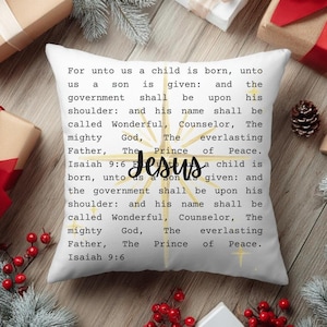 May include: White square pillow with the text "For unto us a child is born..." and the word "Jesus" in cursive. The pillow has a gold starburst design and is surrounded by Christmas decorations.