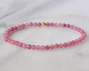 May include: A delicate bracelet featuring small, faceted pink gemstones strung together. A single gold-colored bead is centered on the bracelet. The bracelet is displayed on a white marble surface. Jewelry.