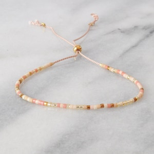May include: A delicate pink and gold beaded bracelet with a sliding knot closure. The bracelet is made with small, round beads in shades of pink and gold, and features a gold bead at the knot.