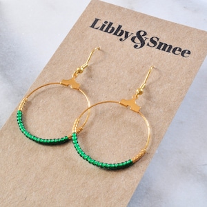 May include: Gold hoop earrings with green beads. The earrings are on a brown card with the text "Libby & Smee" printed on it.
