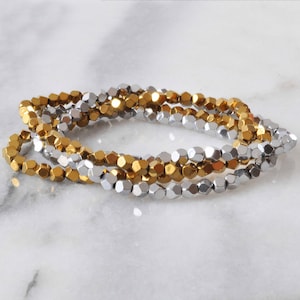 May include: A close-up of a silver and gold beaded bracelet. The beads are faceted and have a hexagonal shape. The bracelet is made of two strands of beads, one silver and one gold.