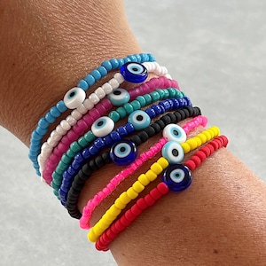 Puede incluir: Una pila de pulseras de cuentas de colores con dijes de ojo azul maligno. Las pulseras están superpuestas en la muñeca de una persona.