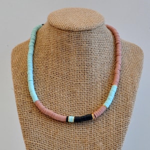 May include: A light blue, pink, and black beaded necklace with gold accents. The necklace is made of round, flat beads and is displayed on a burlap mannequin.