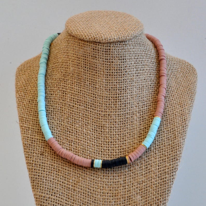 May include: A light blue, pink, and black beaded necklace with gold accents. The necklace is made of round, flat beads and is displayed on a burlap mannequin.