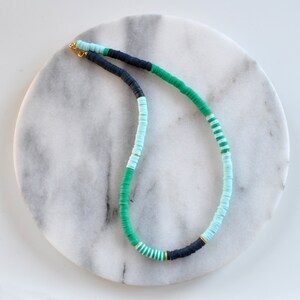 May include: A green, blue, and black beaded necklace with gold accents. The necklace is made of round, flat beads and is displayed on a white marble surface.