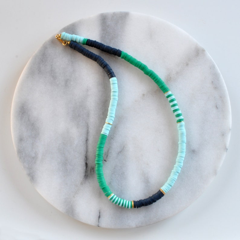 May include: A green, blue, and black beaded necklace with gold accents. The necklace is made of round, flat beads and is displayed on a white marble surface.