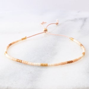 May include: A delicate bracelet with small white and peach beads and gold accents. The bracelet is adjustable with a pink and white cord.