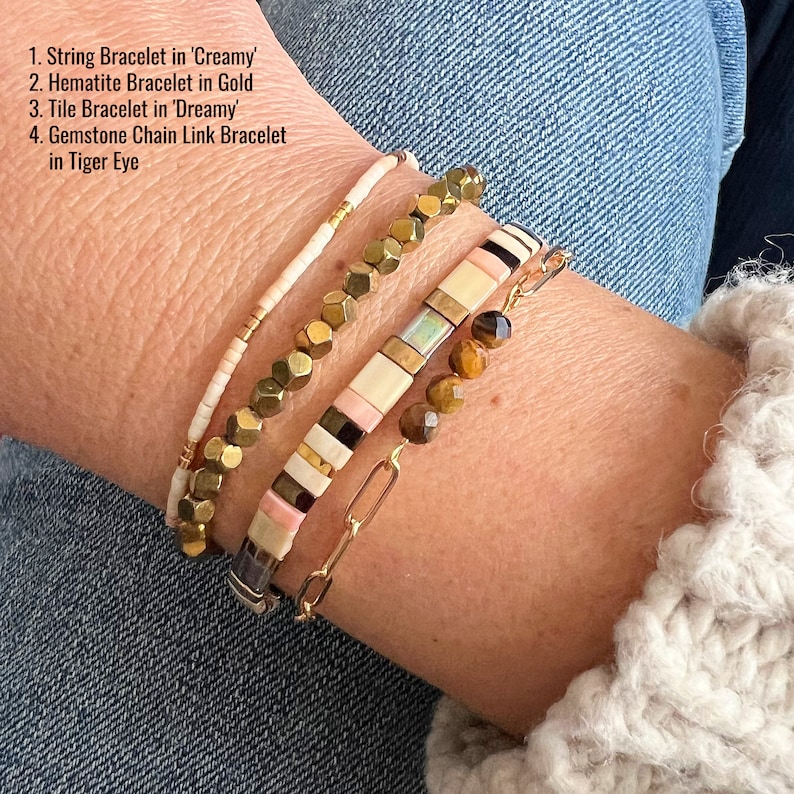 May include: A close-up of a wrist wearing four bracelets. The top bracelet is a thin white string bracelet with gold beads. The second bracelet is made of gold-toned metal beads. The third bracelet is a multi-colored tile bracelet with pink, white, black, and gold beads. The bottom bracelet is a gold chain link bracelet with tiger eye beads.