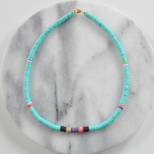 May include: A turquoise blue necklace made of round beads with a colorful stripe detail. The necklace is on a white marble surface.