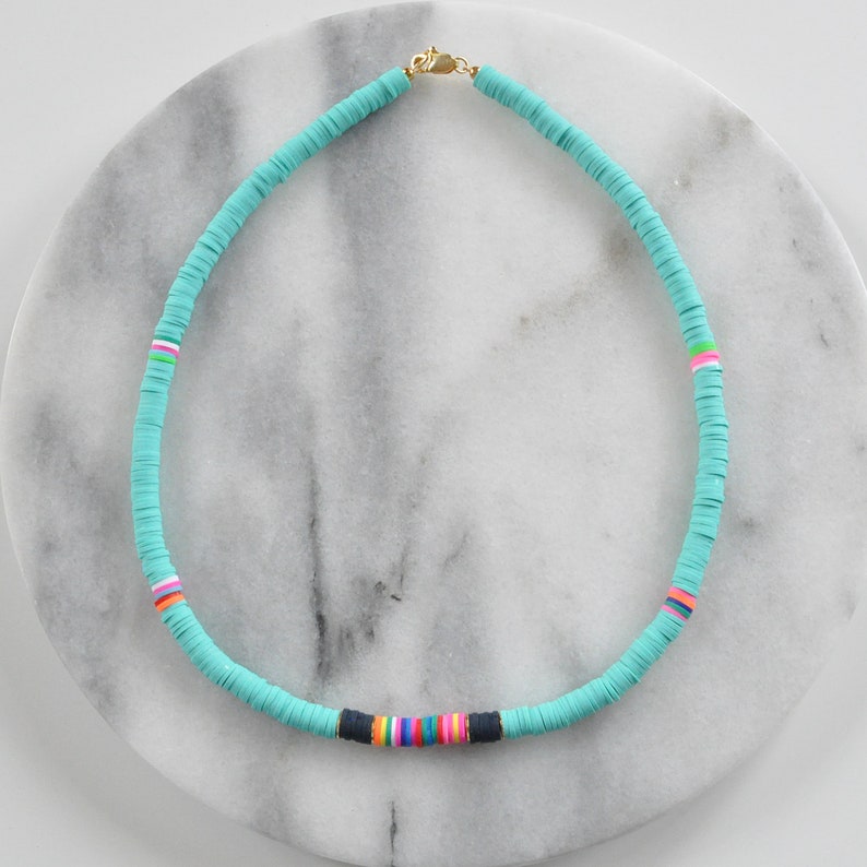 May include: A turquoise blue necklace made of round beads with a colorful stripe detail. The necklace is on a white marble surface.