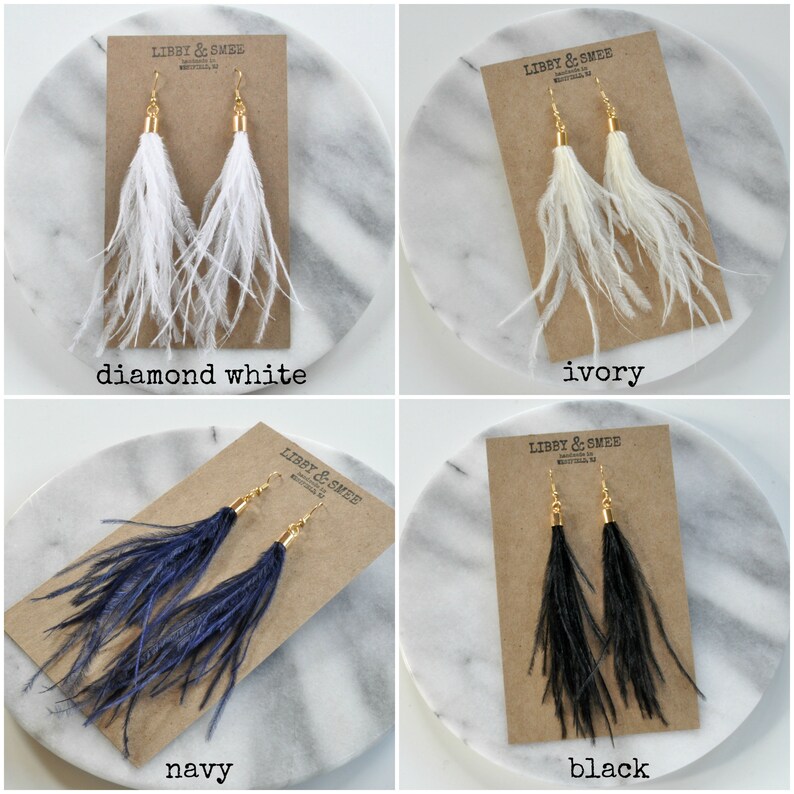 May include: Four pairs of feather earrings on cards with the brand name "Libby & Smee" printed on them. The earrings are in four colors: diamond white, ivory, navy, and black.