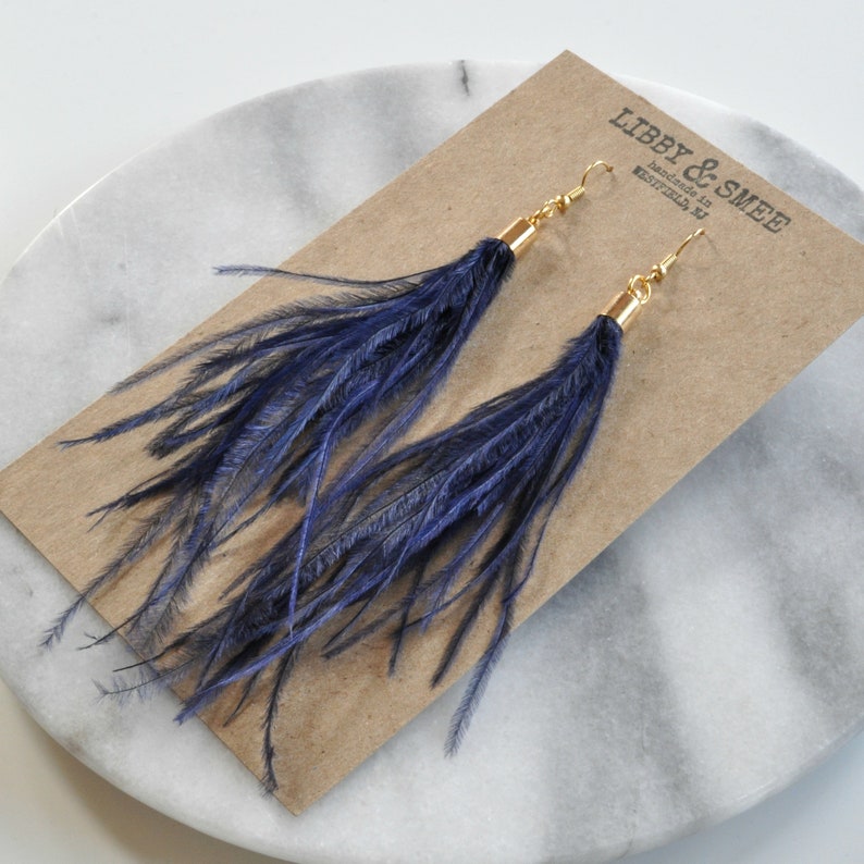 May include: A pair of long, navy blue feather earrings with gold hooks. The earrings are displayed on a brown card with the text "LIBBY & SMEE Handmade in Westfield, NJ".