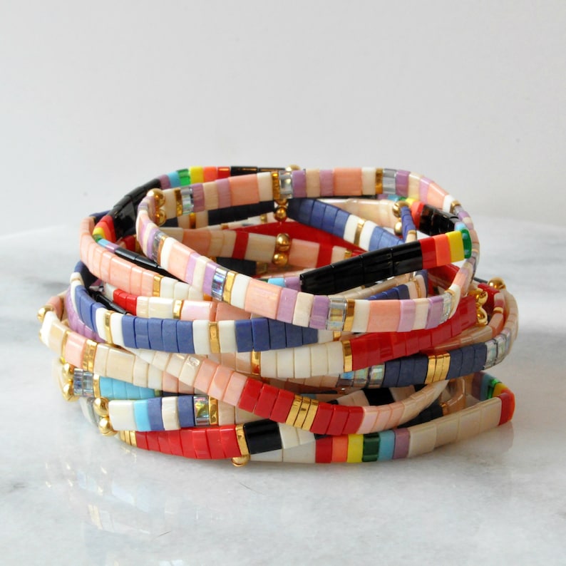 May include: A stack of colorful beaded bracelets in red, blue, pink, white, and black with gold accents. The bracelets are made of small square beads.