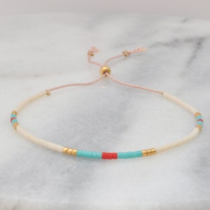 May include: A white beaded bracelet with turquoise and red accents. The bracelet has a gold bead and a pink cord for adjusting the size.