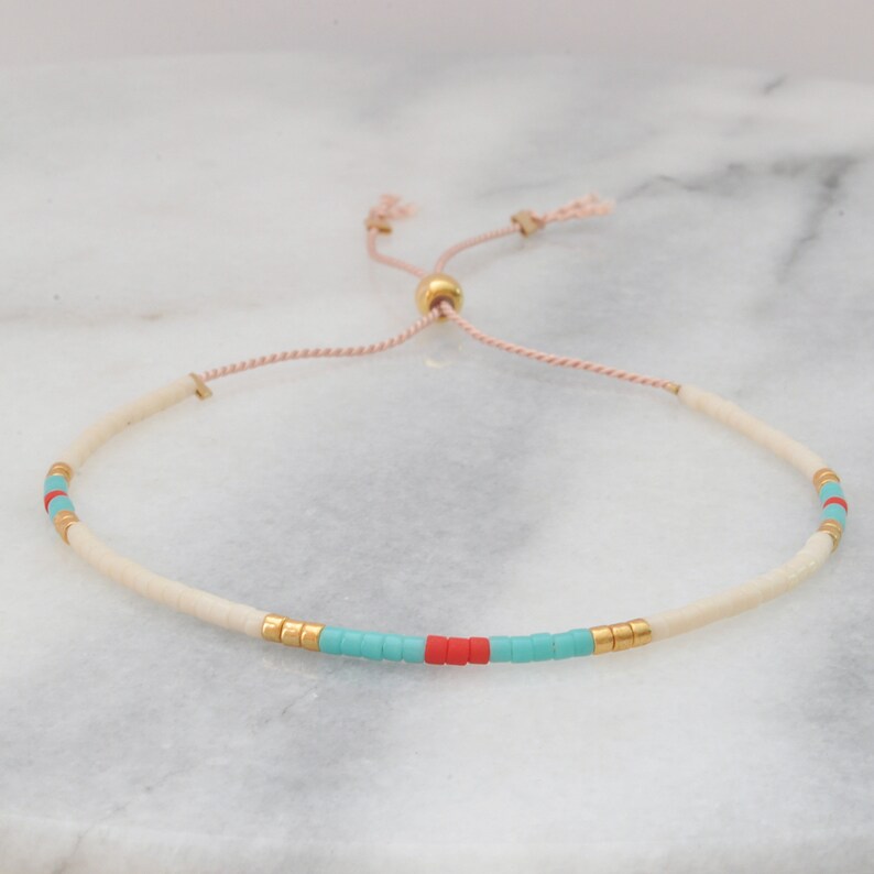 May include: A white beaded bracelet with turquoise and red accents. The bracelet has a gold bead and a pink cord for adjusting the size.