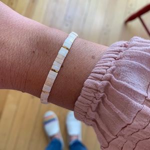 May include: A white and gold beaded bracelet on a person's wrist. The bracelet is made of small, square beads with gold accents.
