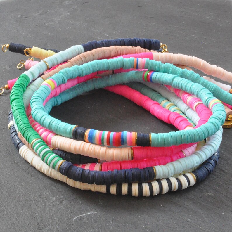 May include: A stack of colorful beaded necklaces made of flat, round, plastic beads. The necklaces are in shades of pink, blue, green, white, and black.