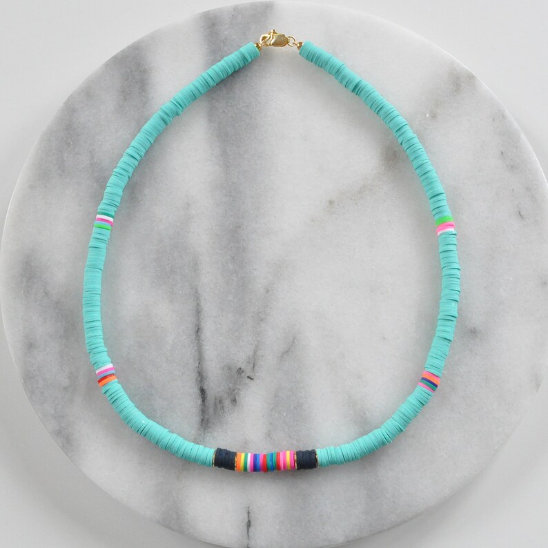 May include: A turquoise blue necklace made of round beads with a colorful stripe detail. The necklace is on a white marble surface.